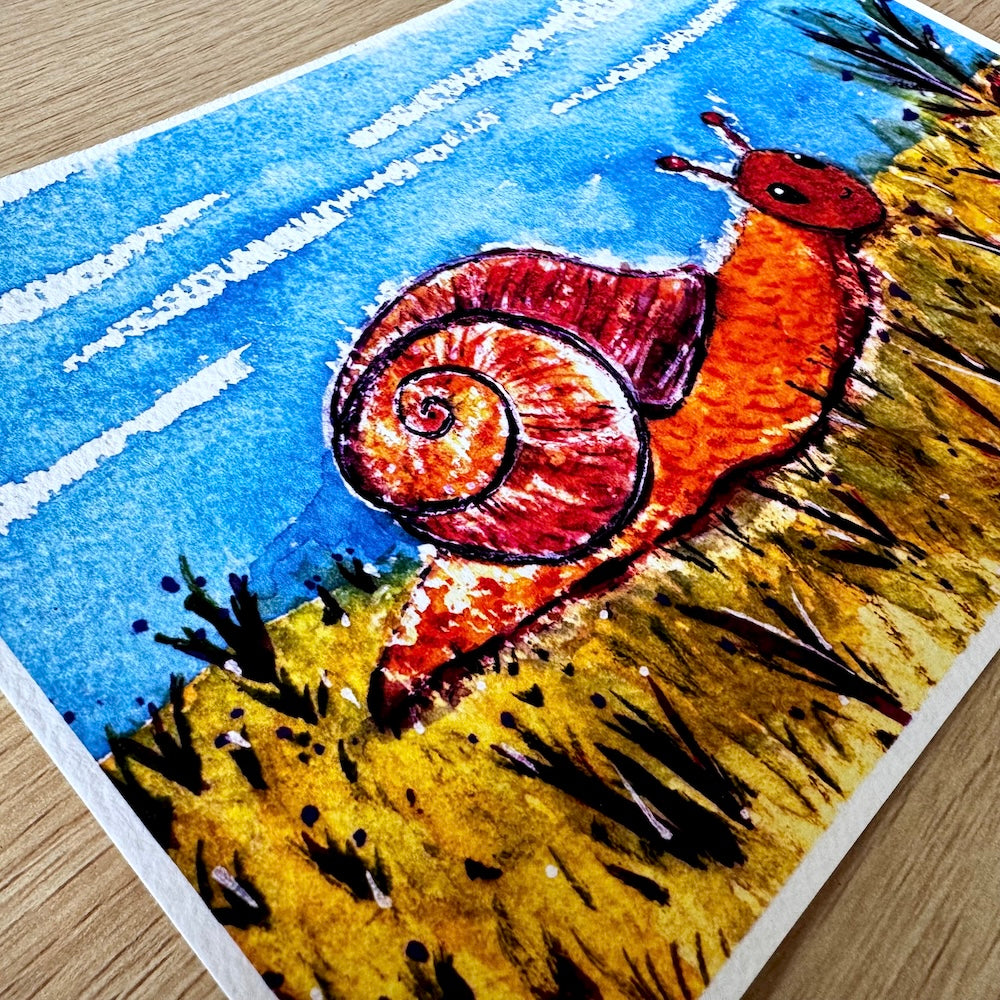 Gros plan d’une illustration aquarelle représentant un escargot orange avec une coquille rouge-brun en spirale, avançant dans une prairie dorée sous un ciel bleu. Les détails de la texture du papier et les couleurs vives donnent vie à cette scène joyeuse et champêtre. Appartient à Boutique de Moments.