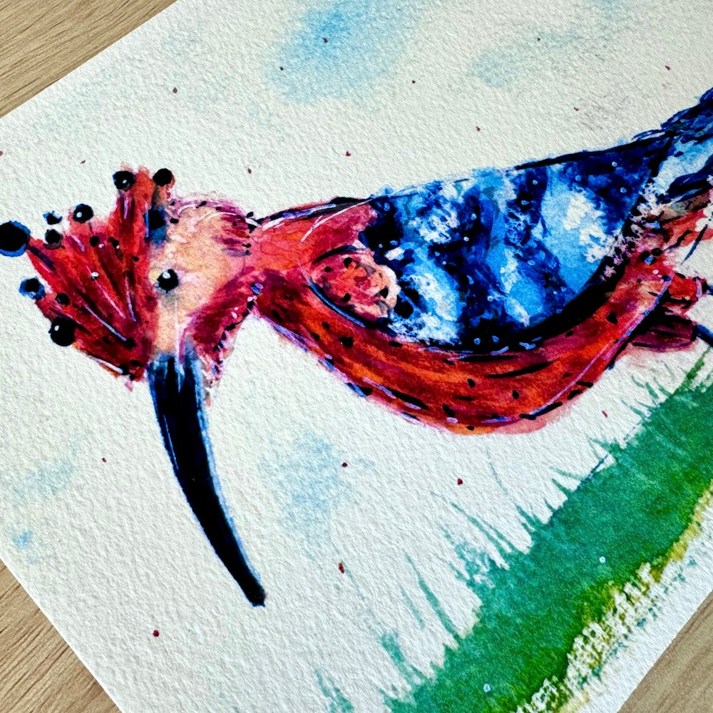 Gros plan d’une illustration aquarelle représentant une huppe fasciée aux couleurs vives, avec un plumage roux et bleu et une crête ornée de pointes noires. La texture du papier est visible, ajoutant du relief et de la profondeur à la scène. Appartient à Boutique de Moments.