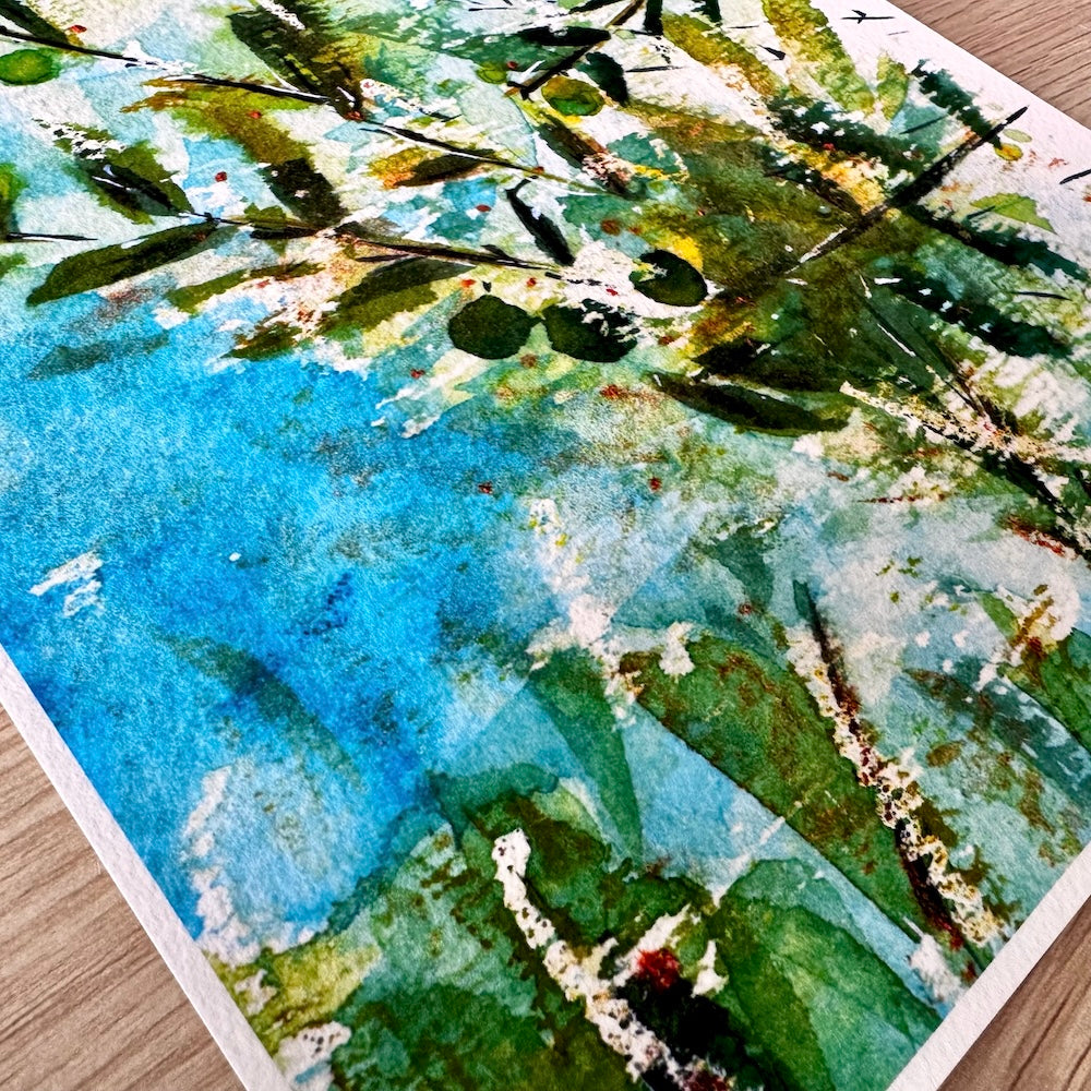 Gros plan d’une illustration aquarelle montrant des rameaux d’olivier aux feuilles vertes et olives naissantes sur un fond bleu lumineux. Les textures délicates du papier mettent en valeur les nuances douces et naturelles de la composition. Appartient à Boutique de Moments.