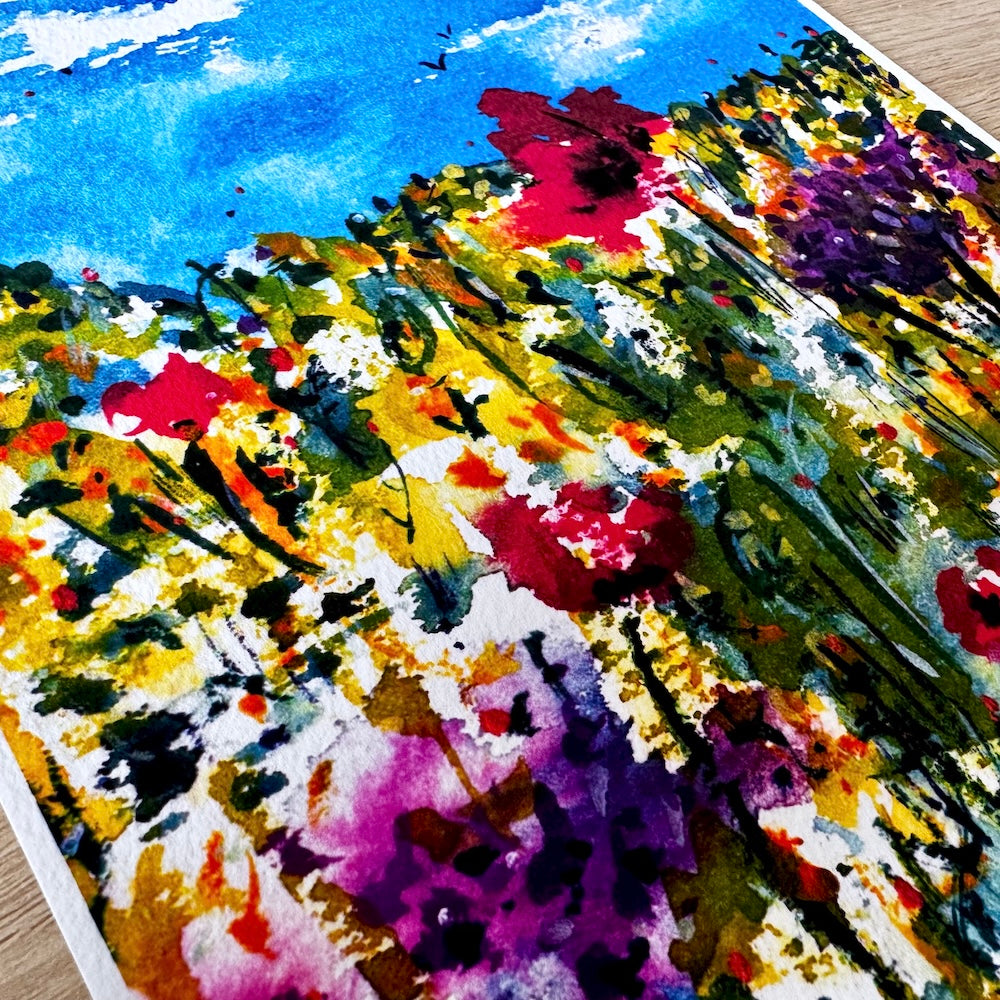 Gros plan d’une illustration aquarelle représentant un champ de fleurs sauvages colorées, mêlant rouges vifs, violets profonds, jaunes lumineux et verts variés, sous un ciel bleu d’été. Les textures du papier et les couleurs éclatantes créent une scène vibrante et joyeuse. Boutique de Moments.